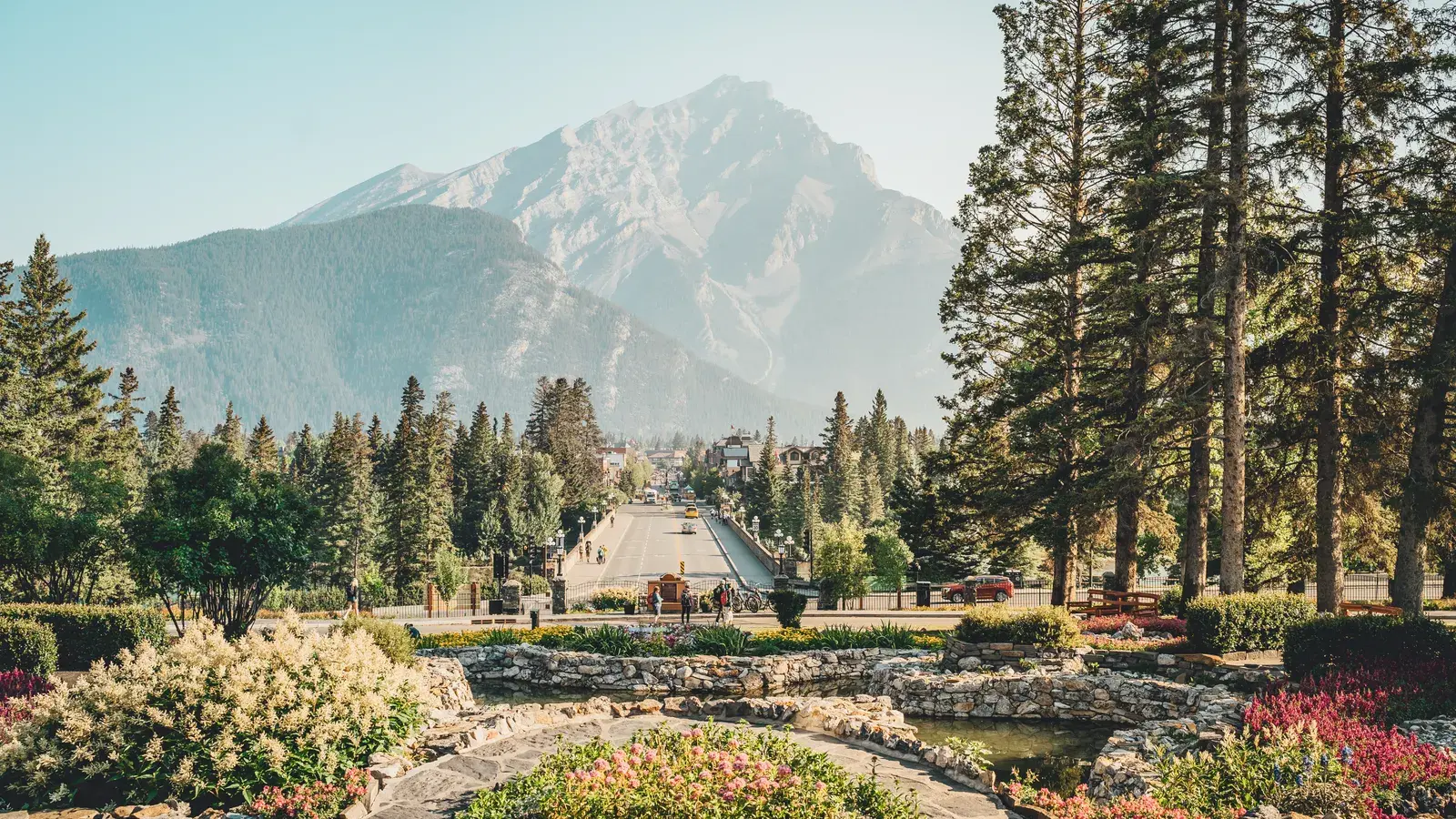 Banff Avenue lined with evergreen trees and flowering shrubs, leading toward a snow-capped mountain peak in the distance