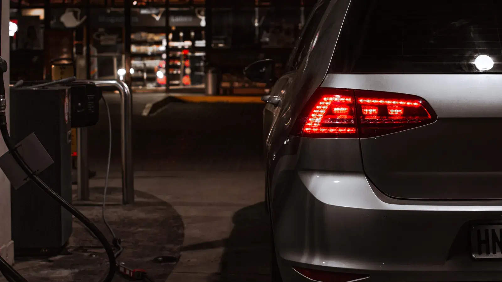 Silver car with illuminated red taillights parked at a nighttime gas station with charging equipment visible