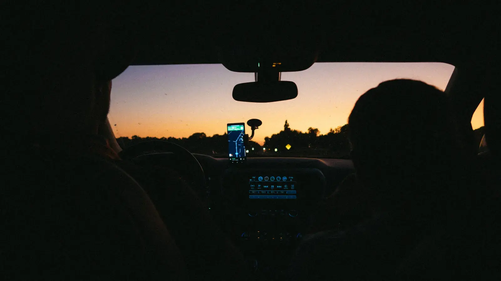 View from inside a car at dusk with a navigation app displayed on the dashboard and windshield showing a road ahead with