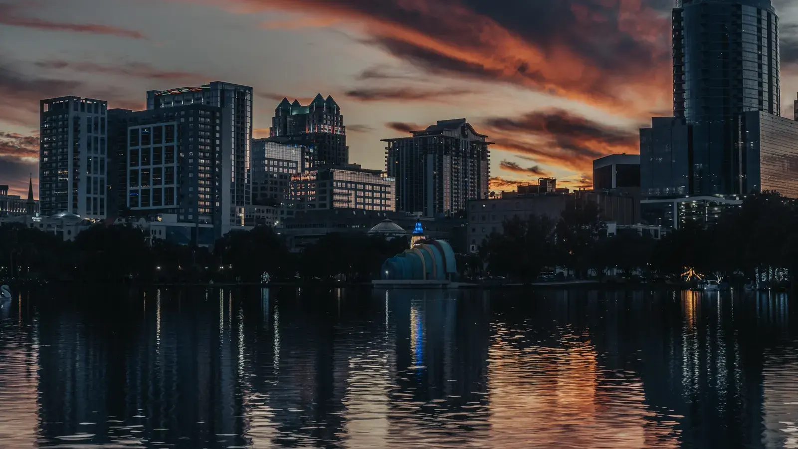 City skyline at dusk with tall buildings reflected in calm water below, orange and blue sky above