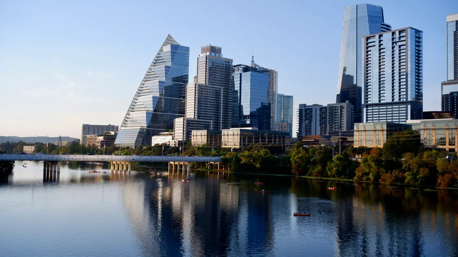 Modern city skyline with tall glass and steel buildings reflected in calm water, with a bridge and green trees along the