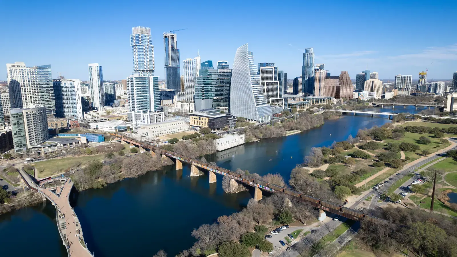Aerial view of a city skyline with tall buildings, a blue river, and a bridge crossing through green parkland
