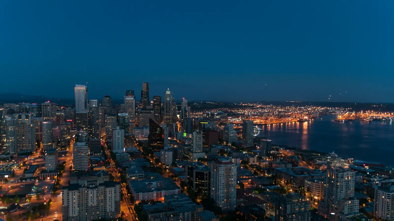 City skyline at twilight with illuminated buildings, streets, and a body of water reflecting lights in the distance