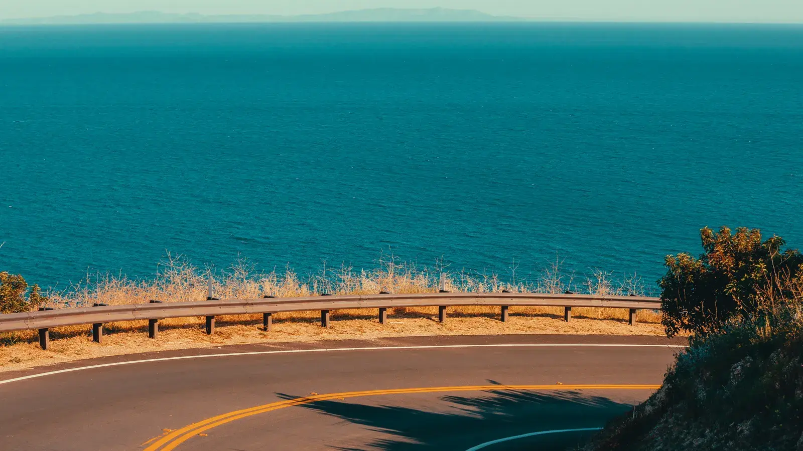 Curved coastal road with yellow center lines overlooking turquoise ocean water and rocky shoreline