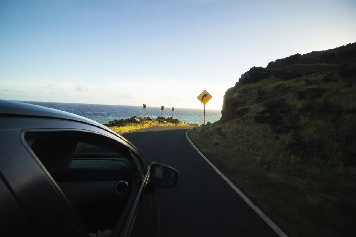 Car driving on a coastal road with ocean view, winding curves, and a yellow warning sign ahead