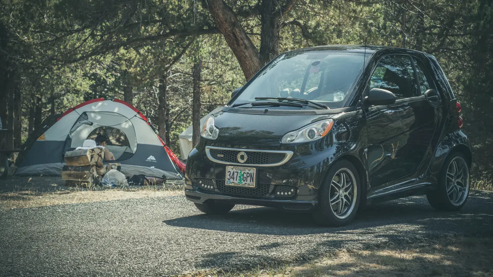 Black compact car parked next to a red and white tent in a wooded campsite