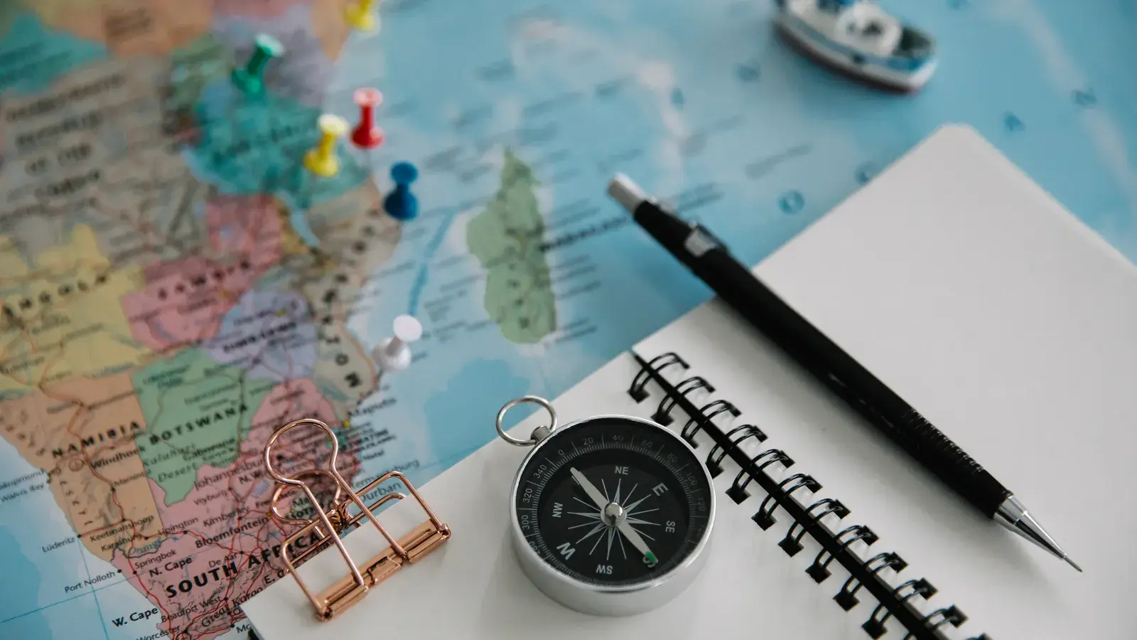 Compass, spiral notebook with pen, and colored pushpins on a map with a toy boat