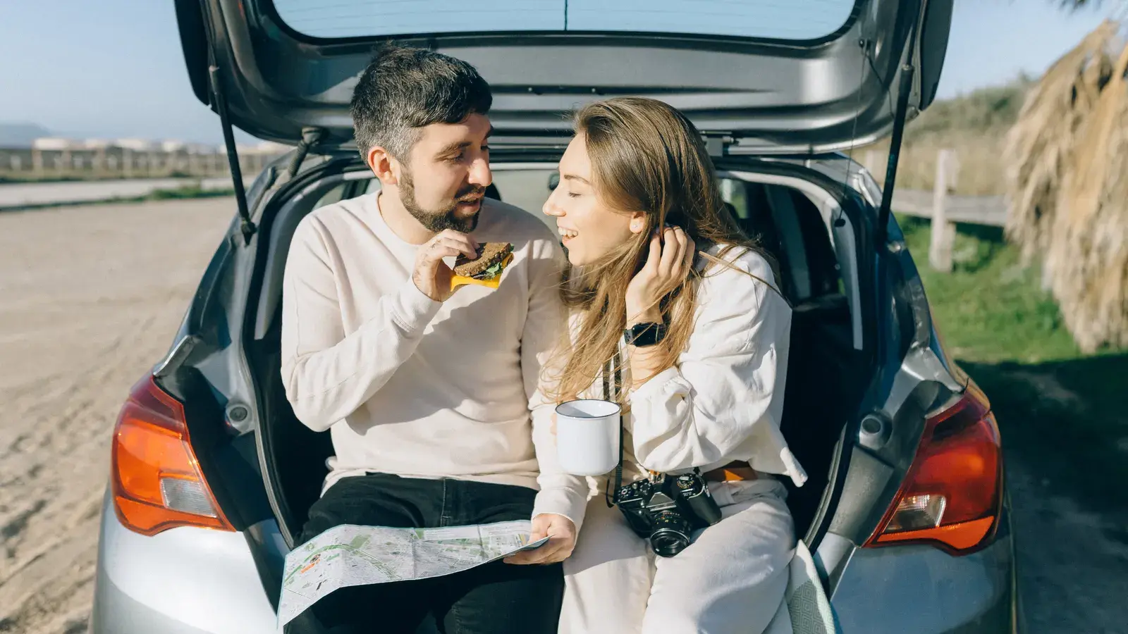 Two people sitting in an open car trunk, eating snacks and looking at a map during a road trip