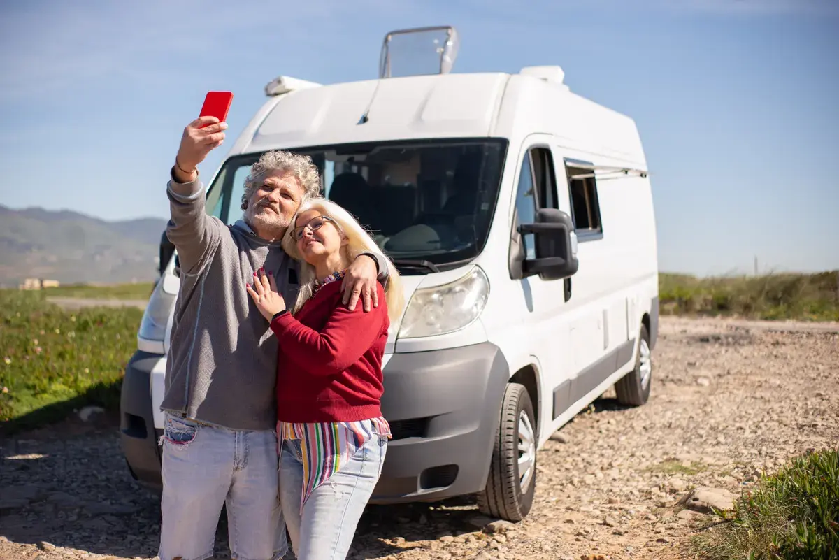 Two people standing in front of a white camper van, holding up a red phone to take a selfie on a rural road