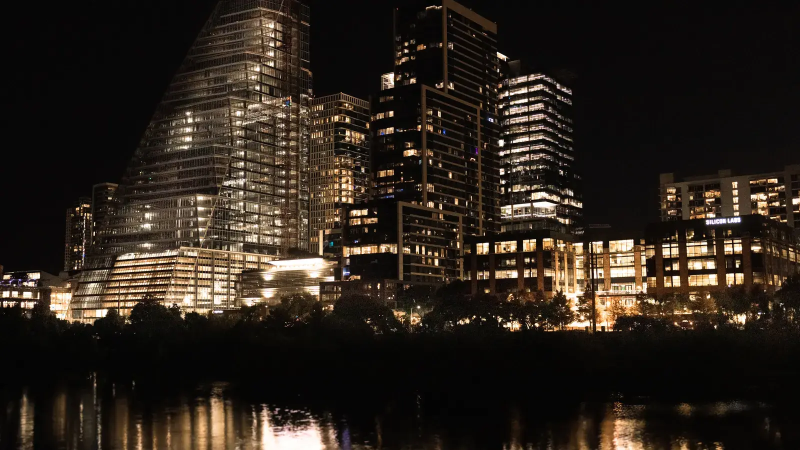Illuminated skyscrapers reflecting in a river at night with trees silhouetted in the foreground