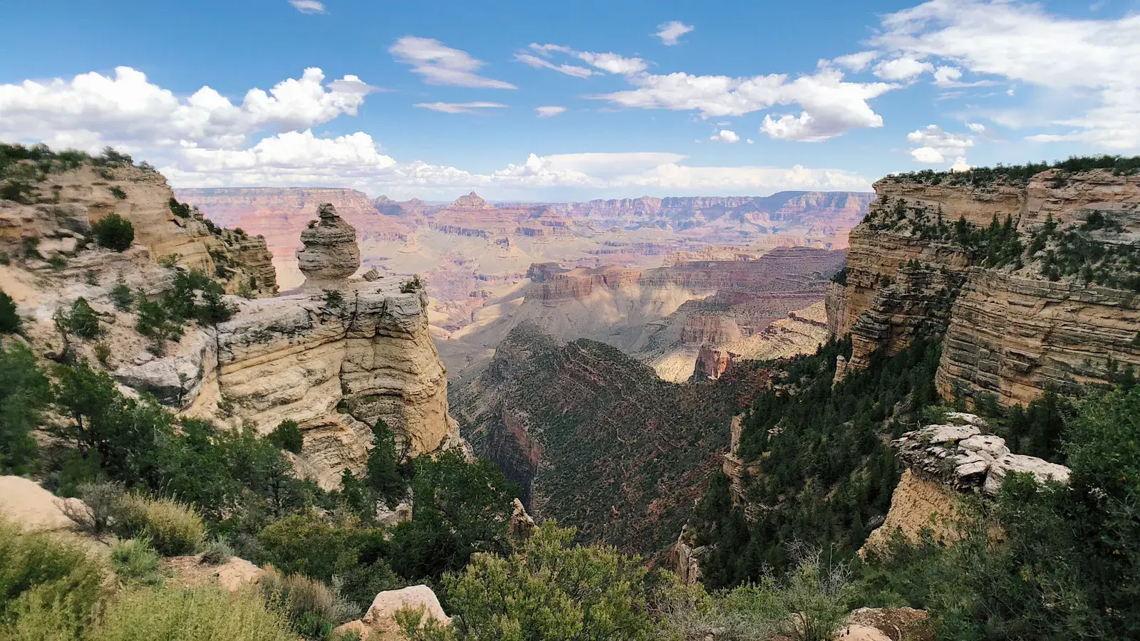 Vast canyon landscape with layered rock formations, deep gorges, scattered vegetation, and blue sky with white clouds