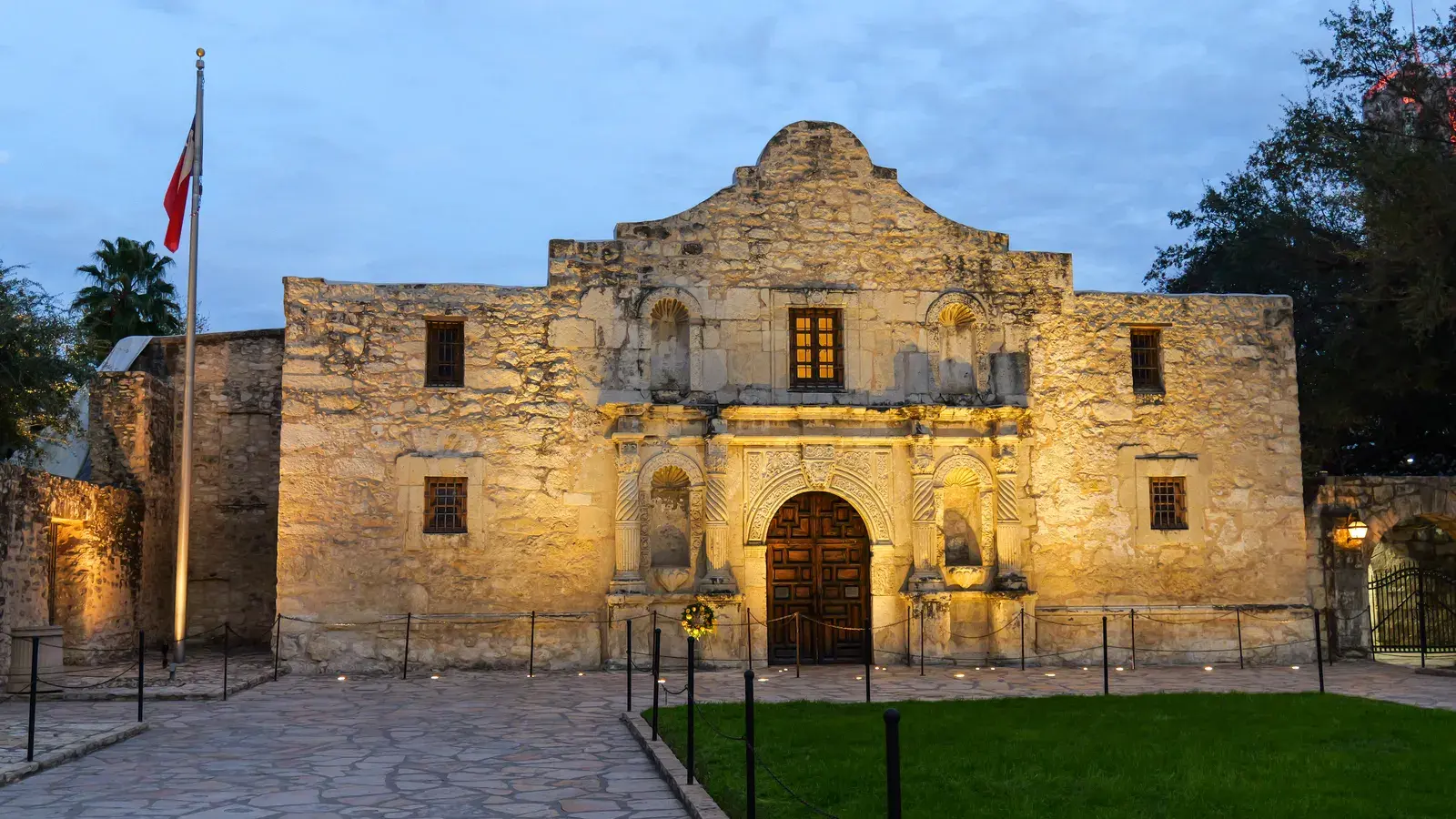 Historic stone building with arched doorway and decorative columns, illuminated at dusk with a flag on the left side