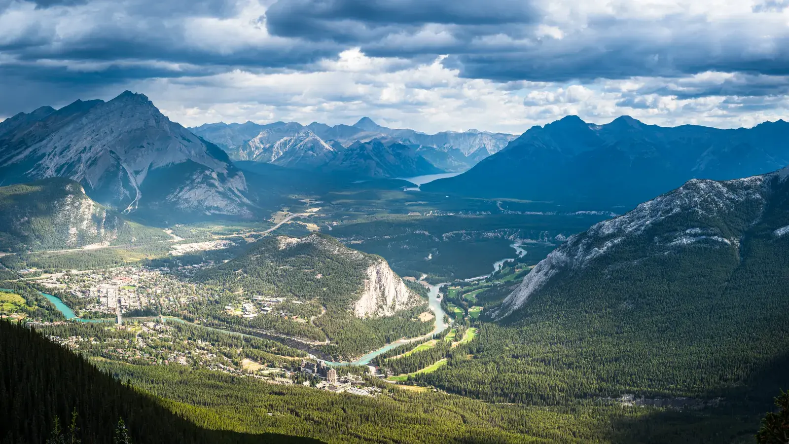 Mountain valley with turquoise river, forested slopes, and snow-capped peaks under cloudy sky