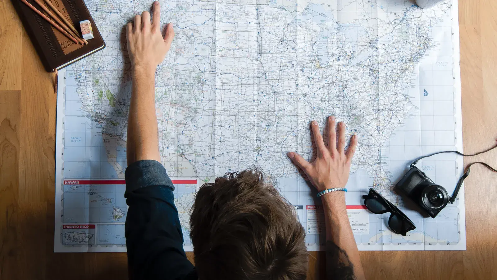 Person pointing at a large paper map on a wooden floor with a camera and travel accessories nearby