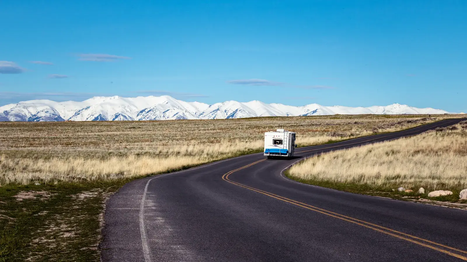 RV traveling on a winding highway through high desert grassland with snow-capped mountains in the distance under clear blue