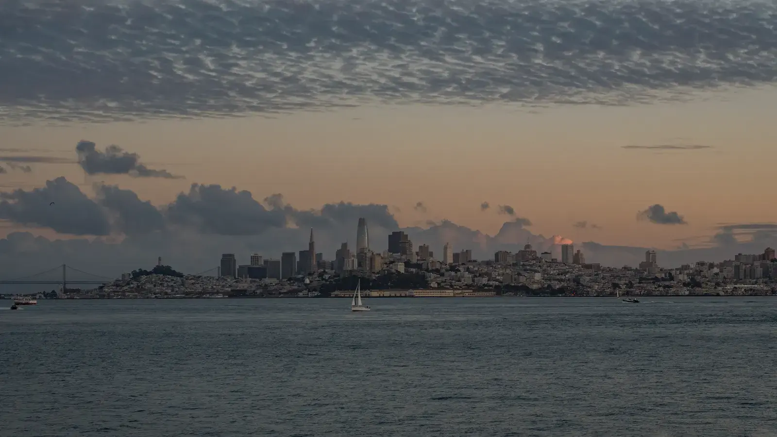 San Francisco skyline at dusk with sailboat on the bay, Golden Gate Bridge visible on the left, and dramatic cloudy sky above