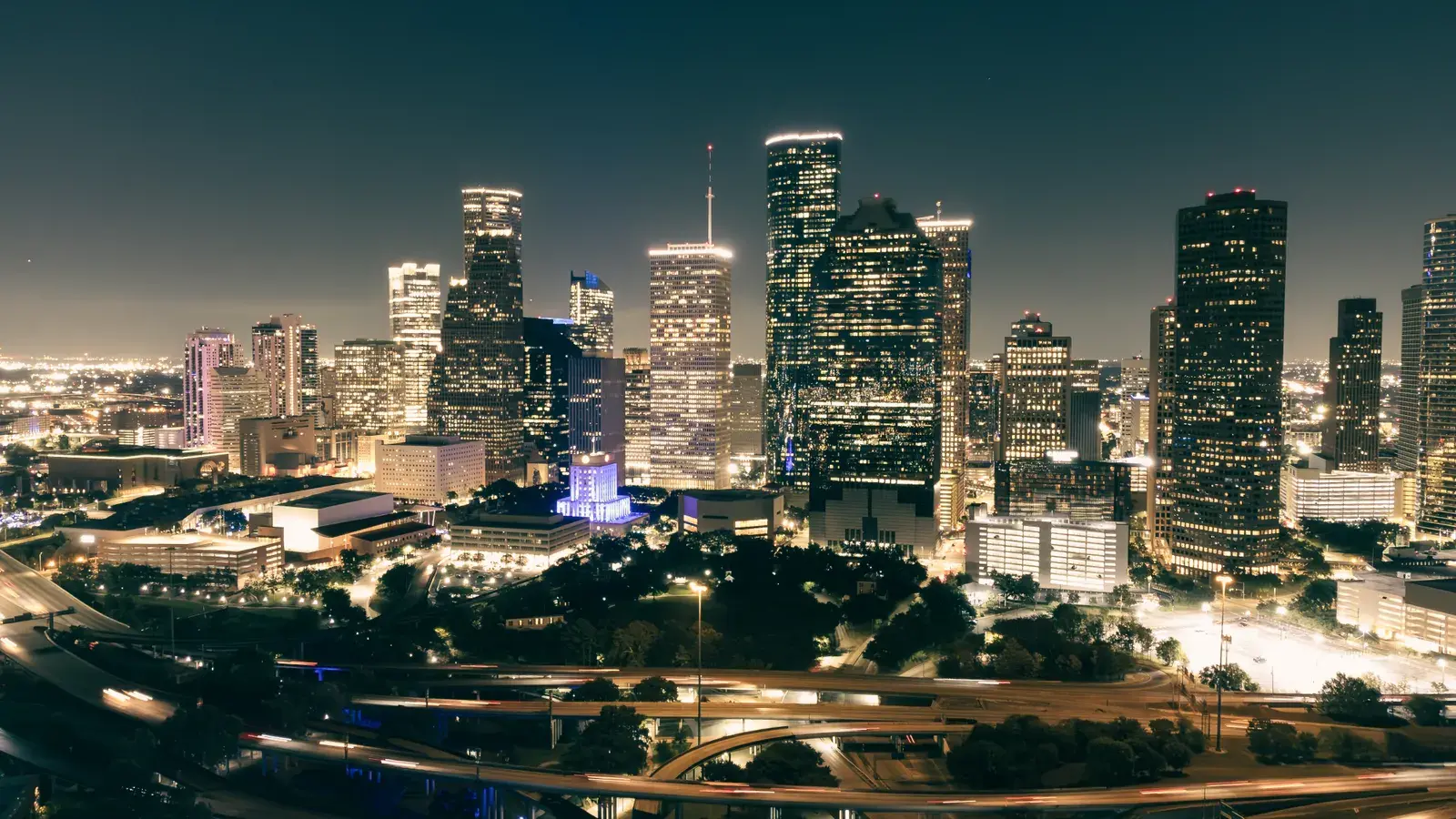 Illuminated downtown skyline at night with tall skyscrapers, highways, and green spaces below
