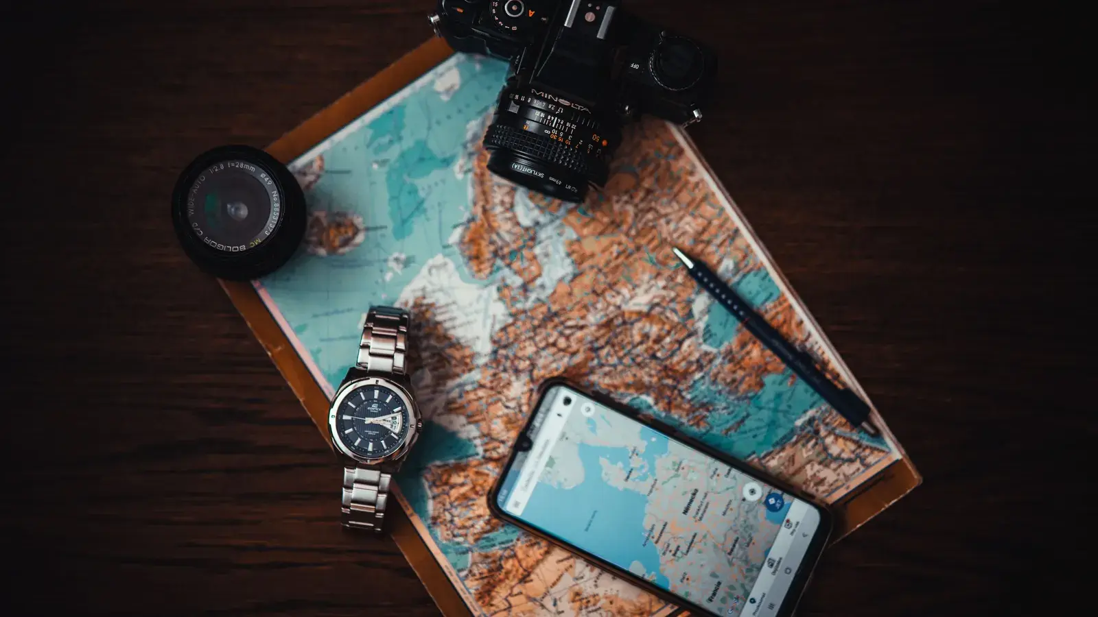 Top view of travel essentials including a map, camera, phone, watch, and lens on a wooden surface.