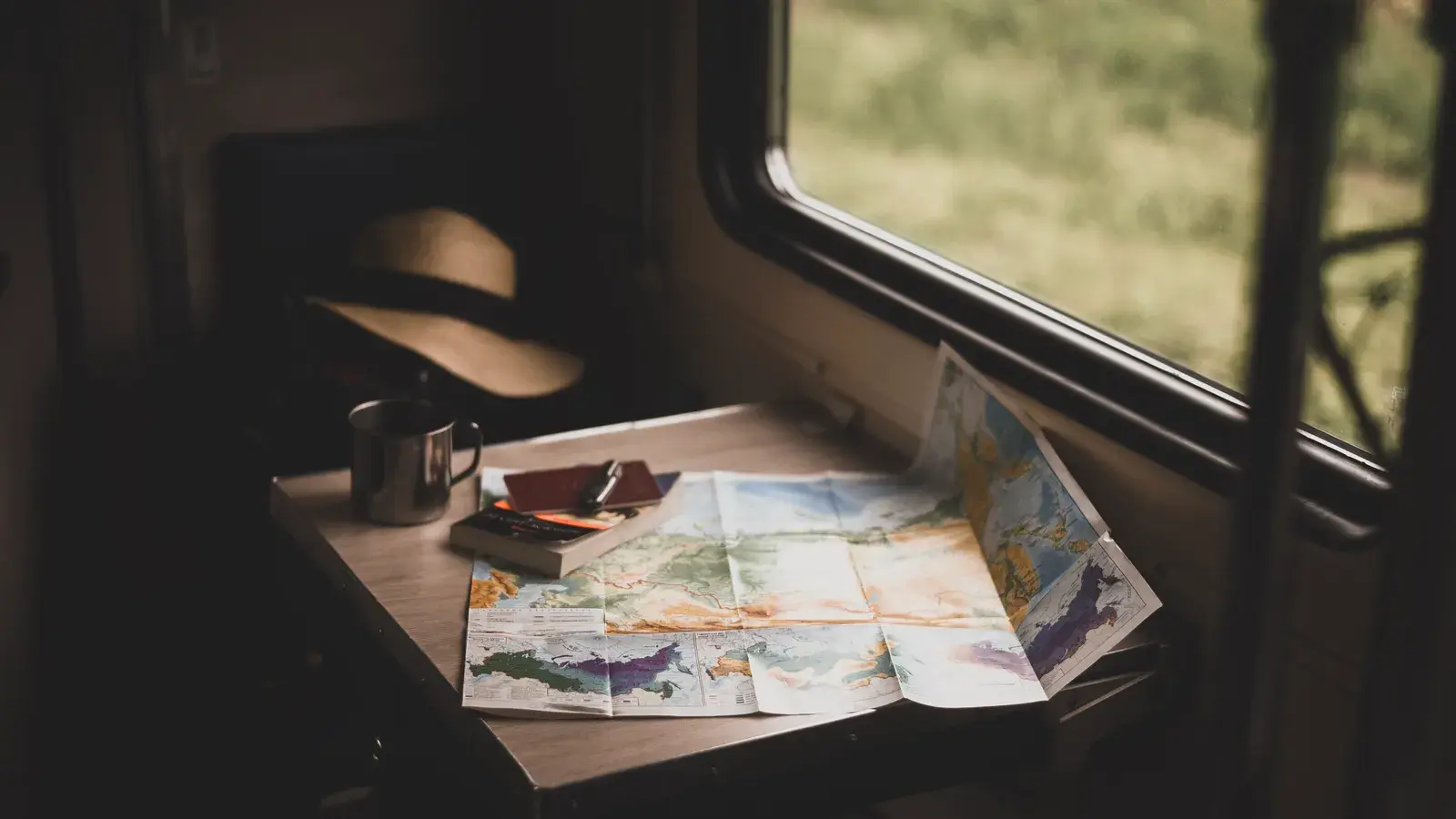 Maps and travel items spread on a table by a train window with countryside visible outside