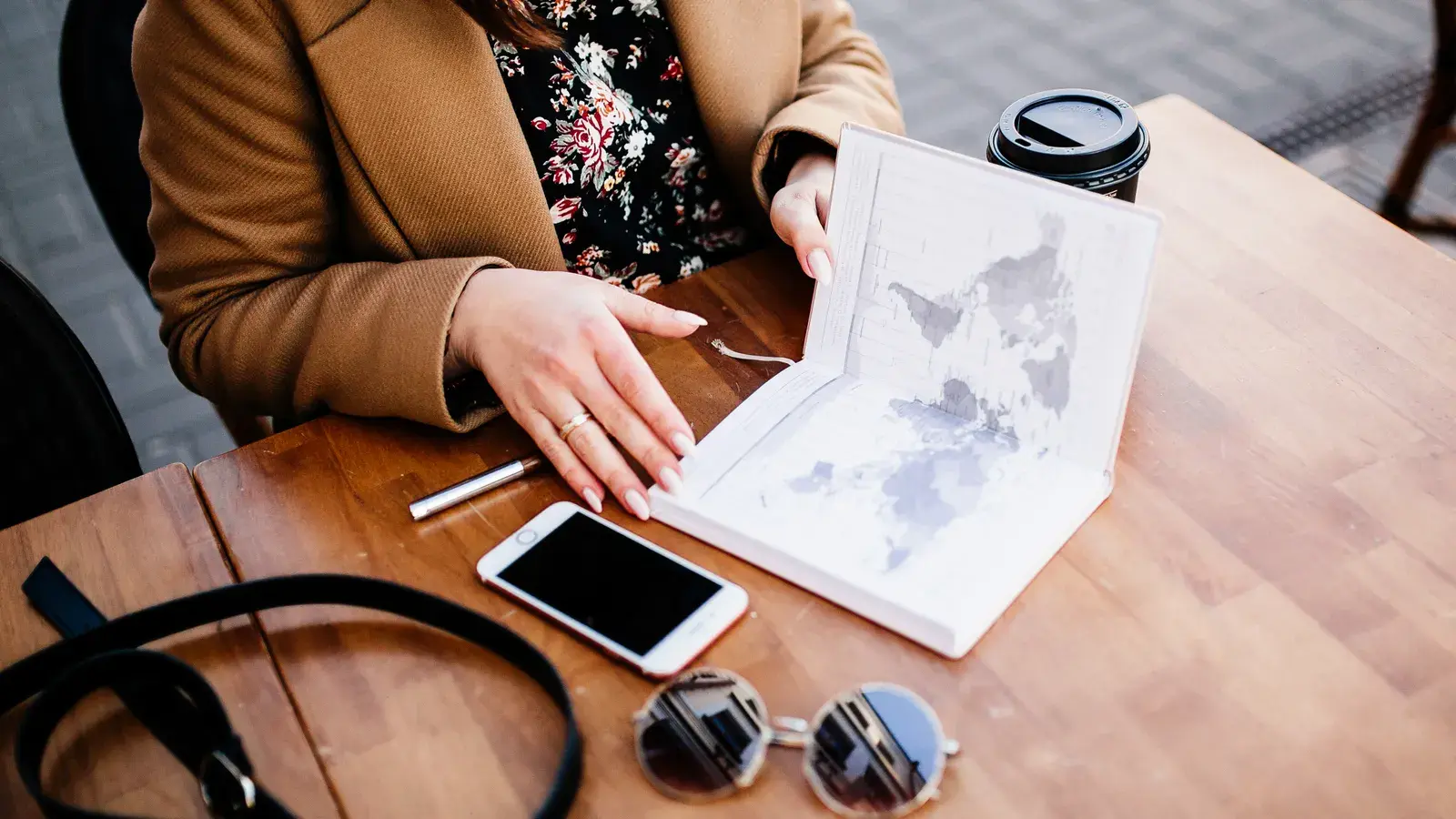 Person in brown jacket reviewing a map at a wooden table with coffee, smartphone, and sunglasses nearby