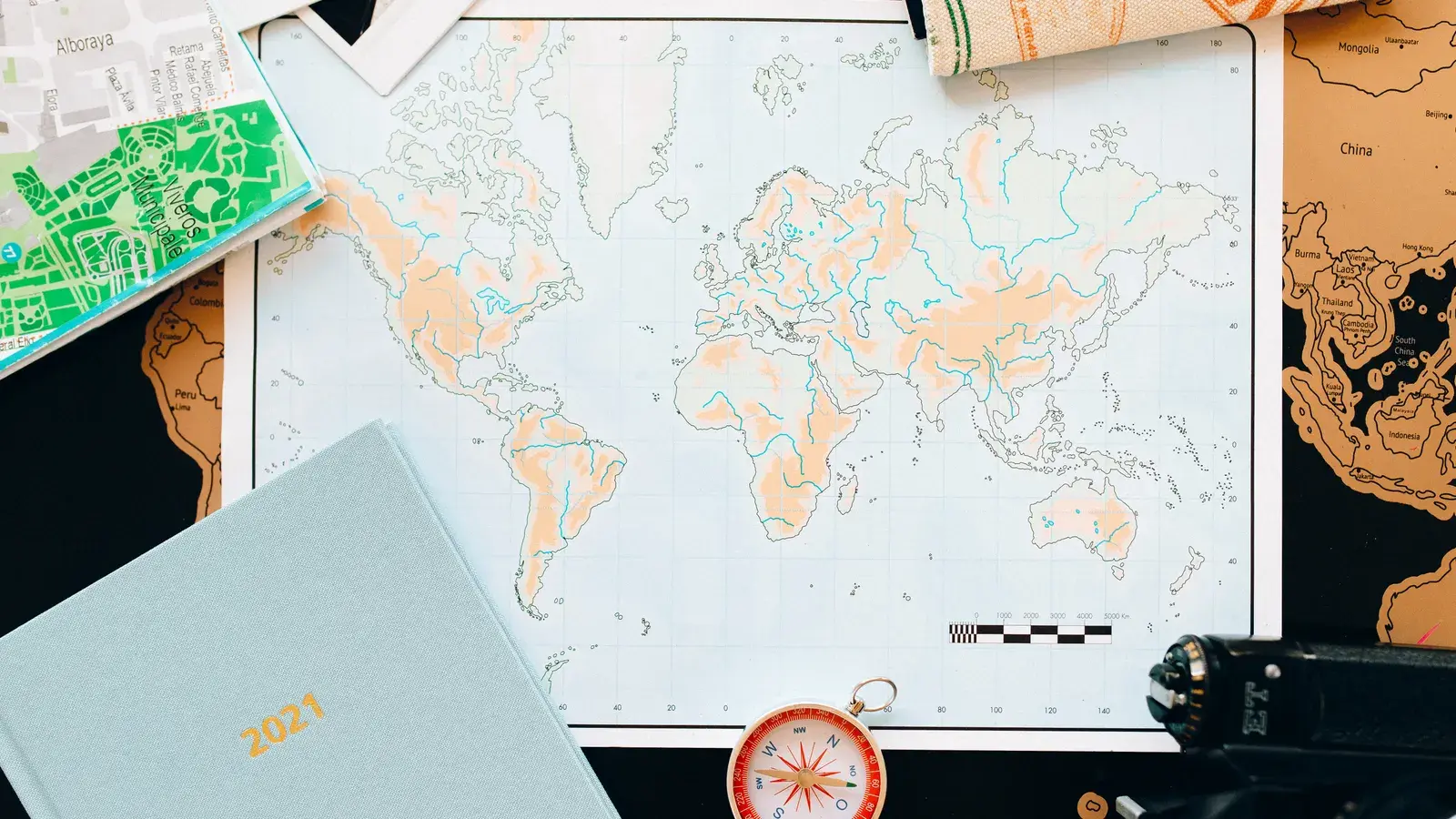 Flat lay of travel planning items including a world map with colored regions, compass, green city map, wooden maps, and
