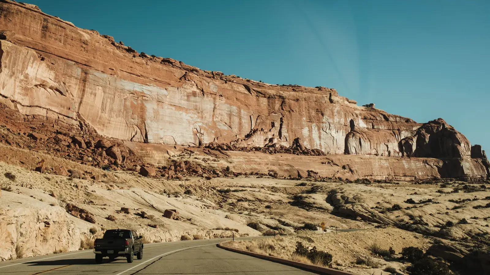 Pickup truck driving on a paved road beneath towering red rock cliffs under clear blue sky
